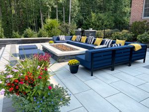 Outdoor patio with modern furniture. A blue sectional sofa with yellow and striped pillows surrounds a rectangular fire pit. Potted plants with colorful flowers are in the foreground, and greenery and trees are in the background. Stone tiles cover the patio floor.
