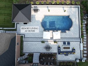 Aerial view of a luxury backyard with a rectangular pool, two large white umbrellas, lounge chairs, a dining area, and a modern fire pit surrounded by seating. A landscaped garden borders the area. Text on the patio reads "Resort Living at Home.
