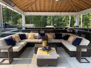 An outdoor patio features a U-shaped wicker sofa with cream cushions and a mix of navy and yellow patterned pillows. A matching wicker ottoman with a gold planter holding flowers and a tray with teacups sits in the center. The area is covered by a wooden pergola.
