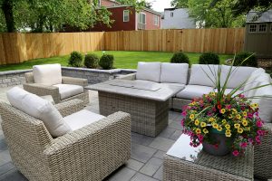 A backyard patio features gray wicker outdoor furniture with beige cushions, including a sofa, two armchairs, and a rectangular fire pit table in the center. A colorful potted plant sits nearby. The area is surrounded by a wooden fence, green lawn, and shrubbery.