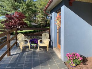 A cozy patio with two yellow Adirondack chairs beside a blue house. The door is decorated with a colorful wreath. There's a flower pot with purple and white flowers between the chairs, and another pot with pink flowers by the wall. Trees and a fence are in the background.